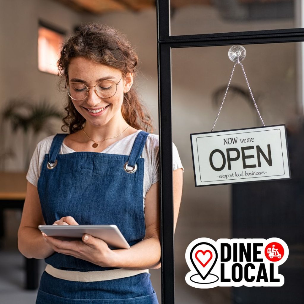 Restaurant owner standing beside an open sign smiling at her tablet that is running the Dine Local online ordering and delivery solution.