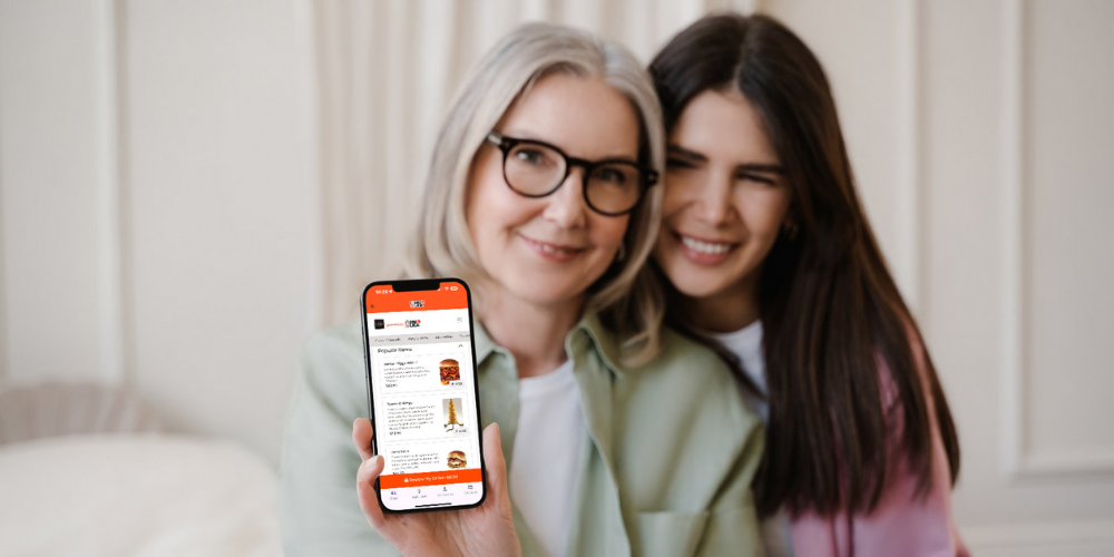 Image of two women, one younger and one older. The older woman is showing her phone with the Dine Local ordering platform on it illustrating that customers of all ages are preferring to order online from a restaurant's own online ordering platform.
